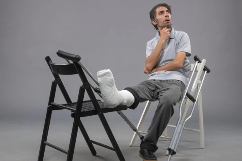 Man with a leg cast and crutches resting his injured leg on a chair while thinking